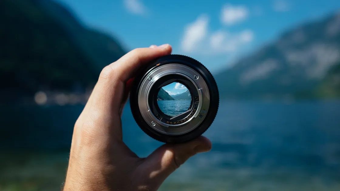 Hand holding a lens with a lake and mountains in the background.